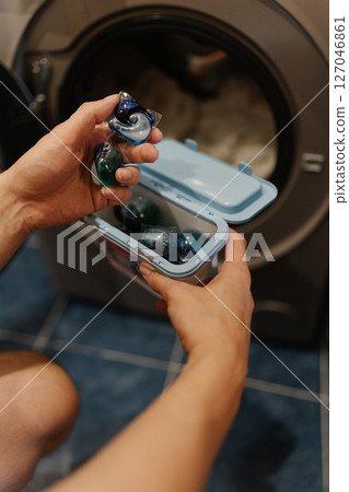 Vertical cropped shot of unrecognizable young man holding two laundry detergent pods preparing to load washing machine with clothes for fresh and clean laundry. Concept of cleanliness and housekeeping 127046861