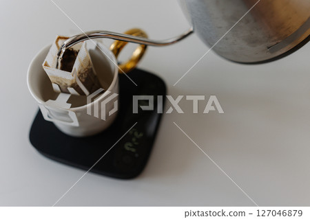 Barista preparing drip coffee using kettle, precision kitchen scales and drip coffee bag, carefully pouring hot water over coffee bag placed in cup on scales, close-up, creating morning ritual. Barista preparing drip coffee using kettle, precision kitchen scales and drip coffee bag, carefully pouring hot water over coffee bag placed in cup on scales, close-up, creating morning ritual. 127046879