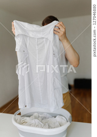 Domestic worker wringing out drenched white t-shirt above laundry basket, wet clothing pooling, preparing laundry for washing machine, vertical shot. Concept of household chores and daily routines. Domestic worker wringing out drenched white t-shirt above laundry basket, wet clothing pooling, preparing laundry for washing machine, vertical shot. Concept of household chores and daily routines. 127046880