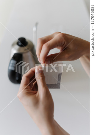 Close-up vertical cropped shot of male hands opening single-serve drip coffee bag, preparing to brew fresh cup of coffee with modern method at home in morning. Process of making freshly brewed coffee. Close-up vertical cropped shot of male hands opening single-serve drip coffee bag, preparing to brew fresh cup of coffee with modern method at home in morning. Process of making freshly brewed coffee. 127046885