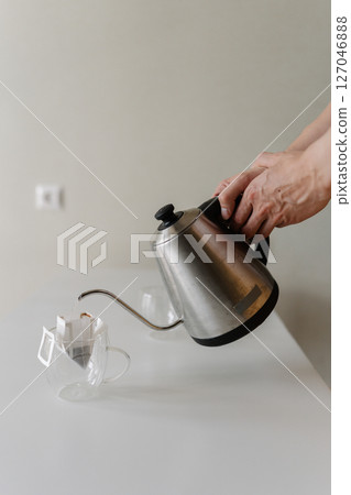 Vertical cropped shot of unrecognizable man preparing drip coffee bag by pouring hot water from stainless steel kettle into clear glass cup placing on white table, creating inviting morning ritual. 127046888