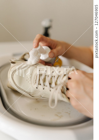 Person applying foam cleaner to dirty white shoe in basin, concentrating on effective shoe care and maintenance while tackling stubborn dirt and grime, close-up. Concept of shoe hygiene. 127046905