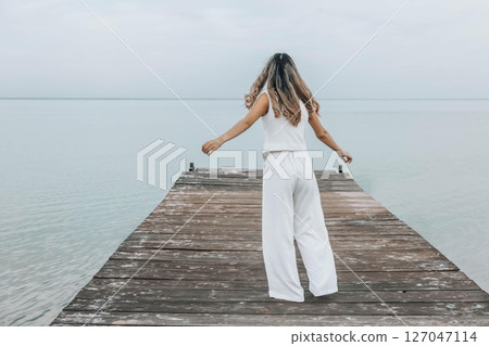 unrecognizable woman dancing barefoot on wooden pier, long hair flowing as she twirls freely, pale sky, calm water, joyful expression in motion, freedom, wellness, motion blur 127047114