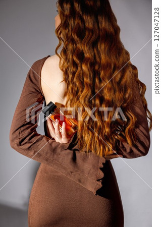 woman in brown dress with long red wavy hair holding perfume bottle behind her back, fashion studio shoot with soft shadows, back view, fragrance branding, beauty marketing, cosmetics retail 127047128
