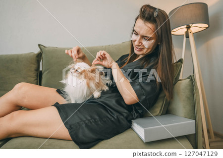 woman in black robe with under eye patches gently petting sleepy japanese chin dog on green sofa in cozy home lamp and houseplant, wellness, pets care, lifestyle brands 127047129
