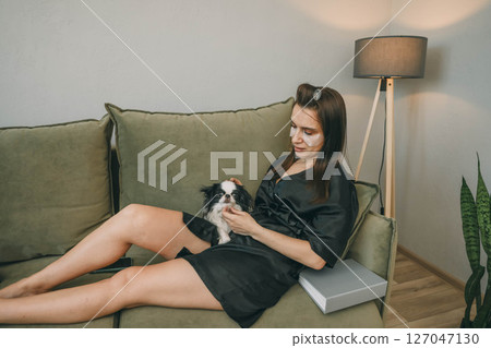 woman with eye patches relaxing on couch with book, and petting dog in cozy living room, concept of self-care and pet care 127047130