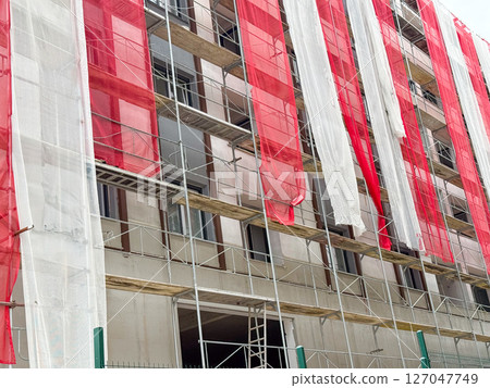 Facade renovation with red and white safety mesh. Urban residential building under exterior construction and scaffolding setup. 127047749
