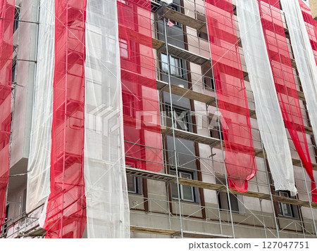 Building under renovation covered with red and white mesh. Urban construction project with scaffolding and protective netting. Building under renovation covered with red and white mesh. Urban construction project with scaffolding and protective netting. 127047751