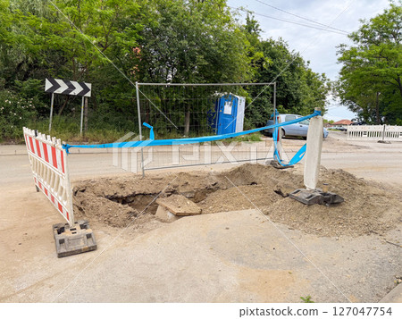 Excavated hole at road construction site with temporary barriers and fencing. Early stage of underground infrastructure repair. 127047754