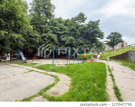 Empty playground with swings and seesaws in a quiet residential area. Peaceful atmosphere of childhood spaces surrounded by summer greenery. Empty playground with swings and seesaws in a quiet residential area. Peaceful atmosphere of childhood spaces surrounded by summer greenery. 127047756
