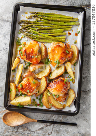 Baked chicken thighs with potato wedges and asparagus close-up on a baking sheet. Vertical top view 127047836