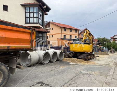 Excavator and concrete pipes at residential roadwork site. Urban infrastructure repair in progress on a suburban street. 127049133