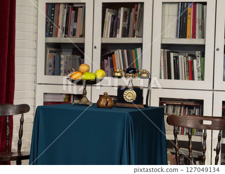 Vintage rotary telephone and fruit bowl on elegant table in front of bookshelf. Classic interior detail in cozy retro-inspired reading room. 127049134