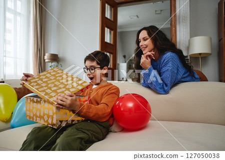 Excited child opens gift with mother nearby in cozy living room setting 127050038