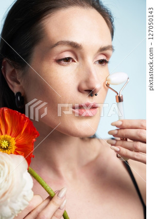 Woman using a facial roller while holding flowers indoors 127050143