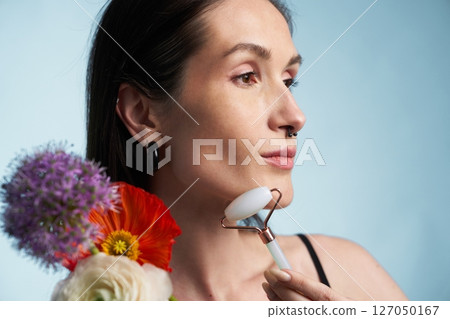 Woman using facial roller with flowers against a light blue background Woman using facial roller with flowers against a light blue background 127050167