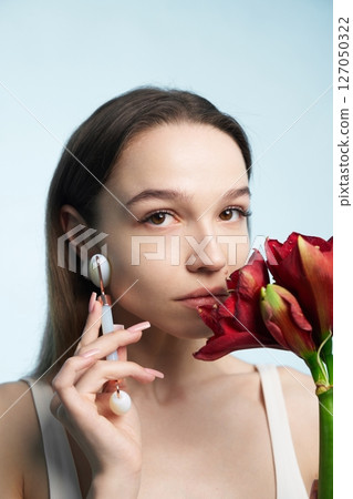 Woman holding red flower while using facial roller in studio setting 127050322
