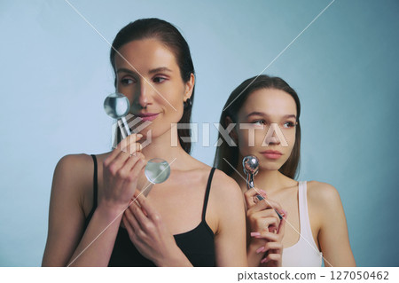 Two women using facial rollers for skincare in a minimalist setting 127050462