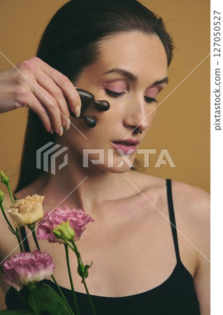 Woman using facial roller on her cheek with flowers in the background 127050527