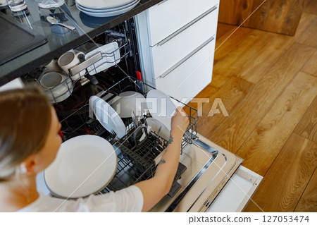 A woman is loading the dishwasher with sparkling clean plates and cutlery in her modern kitchen A woman is loading the dishwasher with sparkling clean plates and cutlery in her modern kitchen 127053474