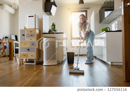 A Charming Woman Happily Engaged in Cleaning the Wooden Floor in a Modern Kitchen Space 127056116