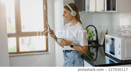 A Young Woman is Happily Enjoying Her Morning Coffee and Listening to Music in a Modern Kitchen 127056117
