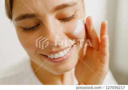 A woman is applying a skincare cream with a bright smile while basking in natural light 127056217