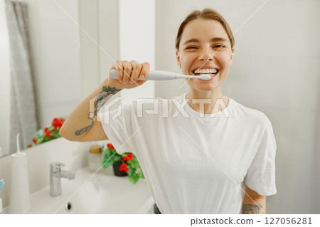 A cheerful woman with a bright smile is brushing her teeth in a modern and stylish bathroom 127056281