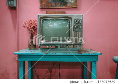 Retro television on a turquoise table against a bright pink wall, with a vase of dried flowers, creating a nostalgic atmosphere in a charming interior setting Retro television on a turquoise table against a bright pink wall, with a vase of dried flowers, creating a nostalgic atmosphere in a charming interior setting 127057629