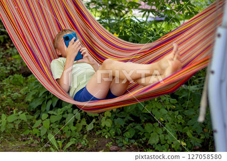 Young boy lies comfortably in colorful hammock while focused on smartphone. Surrounded by lush greenery, he enjoys peaceful, tech-driven outdoor relaxation moment 127058580