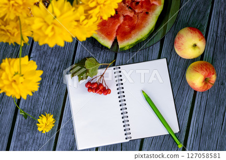 Autumn still life with an open notebook surrounded by bright yellow flowers, apples, juicy watermelon, and red berries on wooden table. For fall-themed projects Autumn still life with an open notebook surrounded by bright yellow flowers, apples, juicy watermelon, and red berries on wooden table. For fall-themed projects 127058581