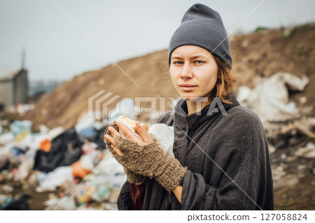 A young homeless woman holds bread in her hands against the background of a garbage dump A young homeless woman holds bread in her hands against the background of a garbage dump 127058824