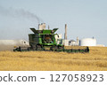 Combine harvester harvesting canola with factory in background 127058923