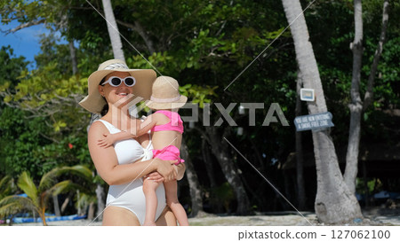 Mom strolls along beach with baby daughter under palm trees enjoying summer vacation 127062100