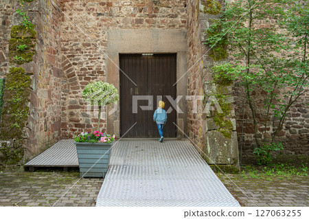 Young child wearing a yellow hat and blue jacket stands in front of a large wooden door set in a rustic stone wall, surrounded by greenery and potted plants on a cobblestone path. 127063255