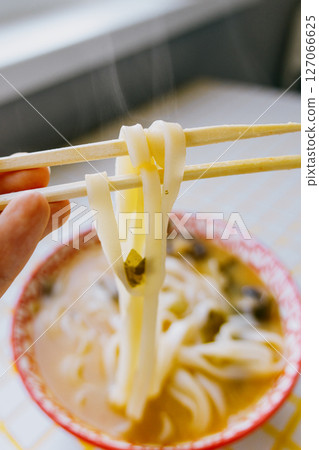 person is eating noodles with chopsticks. colorful bright red plate of ramen sits on yellow table. person is eating noodles with chopsticks. colorful bright red plate of ramen sits on yellow table. 127066625