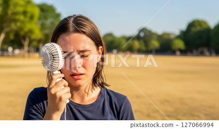 Young woman cooling off with handheld fan in hot weather at outdoor park during summer 127066994