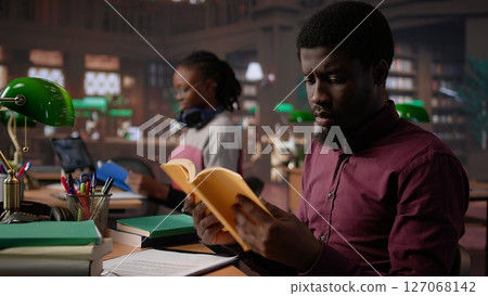 African american guy studies for a critical law exam and reading specialty books to complete class notes with reliable academic information. Studying in the library before the test. Camera B. 127068142