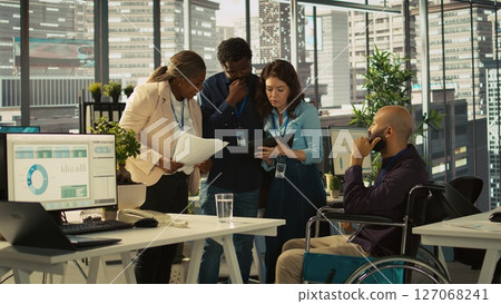 Employees and coworker with disability looking over revenue figures in inclusive office. Man in wheelchair working together with colleagues, comparing company statistical numbers, camera B Employees and coworker with disability looking over revenue figures in inclusive office. Man in wheelchair working together with colleagues, comparing company statistical numbers, camera B 127068241