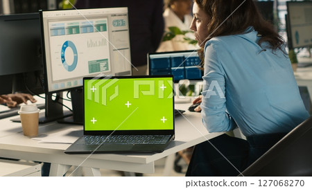 Green screen laptop on office desk next to employee editing documents on computer. Mockup notebook next to worker analyzing data charts, interpreting statistical information, camera A 127068270