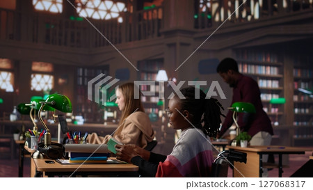Girl pushing her colleague with physical impairment through the library, studying for a new exam with academic materials from the campus database. African american student in wheelchair. Camera A. Girl pushing her colleague with physical impairment through the library, studying for a new exam with academic materials from the campus database. African american student in wheelchair. Camera A. 127068317
