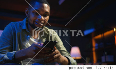 Casual african american man browsing on his smartphone in a comfortable space, sitting at the coffee table. Modern home highlights leisure and online connectivity in a stylish loft. Camera A. Casual african american man browsing on his smartphone in a comfortable space, sitting at the coffee table. Modern home highlights leisure and online connectivity in a stylish loft. Camera A. 127068412