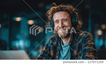 Man wearing headphones smiling while working on laptop in cozy indoor setting with warm lighting and casual style 127071284