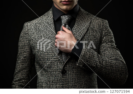 Stylish man in patterned suit adjusts his tie, showcasing modern fashion against dark background Stylish man in patterned suit adjusts his tie, showcasing modern fashion against dark background 127071594
