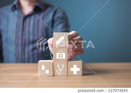 A person arranging wooden blocks with healthcare icons including a syringe, stethoscope, cross, and medication, symbolizing medical care, health services, and wellness solutions on a blue background. A person arranging wooden blocks with healthcare icons including a syringe, stethoscope, cross, and medication, symbolizing medical care, health services, and wellness solutions on a blue background. 127074845