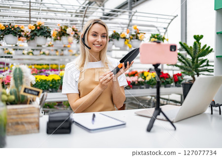 A Caucasian woman in a florist shop demonstrates a gardening tool, engaging in a live video session with flowers in the background. 127077734