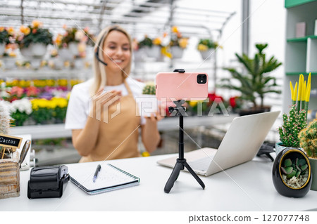 A Caucasian florist broadcasts live from her shop. She is selling a cactus from her collection of plants. 127077748