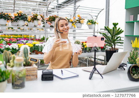 A Caucasian woman in a florist shop presents a cactus while live streaming for online sale from her table. A Caucasian woman in a florist shop presents a cactus while live streaming for online sale from her table. 127077766