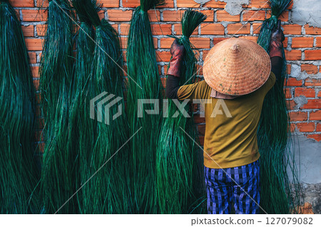 Vietnamese woman in traditional straw hat dries dyed green reeds at a woven mat factory in a village in Vietnam 127079082