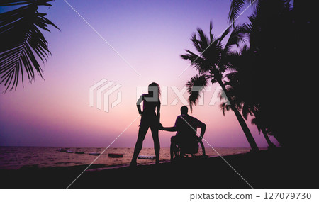 Disabled man in a wheelchair with his wife on the beach. Silhouettes at sunset 127079730